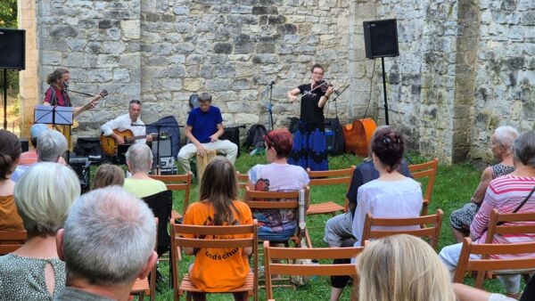 Eine Band, Gittarist, Bassist, Trommer, Geigerin vor alten Mauern. Davor sind Holz-Klappstühle aufgereiht, auf denen die Zuschauer sitzen. Das Publikum ist gemischt von jung bis alt.