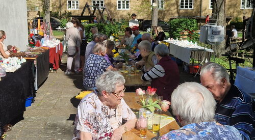 Biertischgarnituren, draußen, bei Sonne, Menschen sitzen an mit Getränken an Tischen. Rechts an der Wand eines Gebäudes sind Tische mit Deko zum Verkauf.