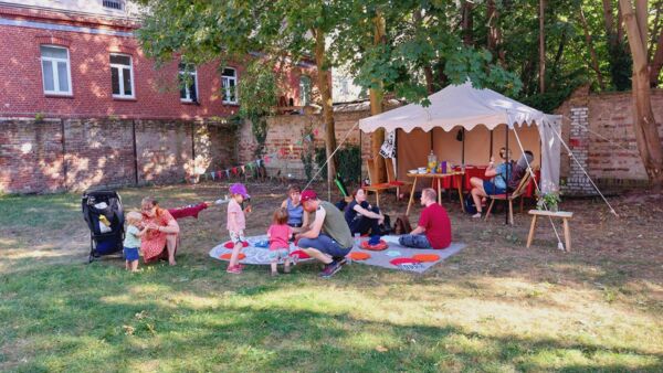 Draußen, Sonne scheint. Picknickdecken auf einer Wiese, eine Art offenes Zelt mit Tisch und Stühlen. Ein paar Erwachsene und kleie Kinder auf der Wiese und den Picknickdecken.