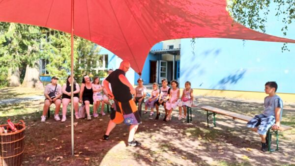 Draußen, Sonne scheint. Großes rotes Sonnensegel. Ein paar Kinder und zwei Frauen sitzen auf Bänken und sehen einem Mann mit Glatze beim Tanzen zu.