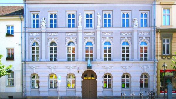 Das Gebäude von außen, von der Straße aus. Das Gebäude und seine Fassade ist aufwendig mit römischen Soldatenfiguren und den beiden Kriegsgöttern Mars und Bellona geschmückt. Es hat viele Fenster mit weißen Rahmen. Die Fassade ist bläulich.