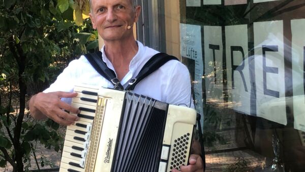 Ein älterer Mann mit kurzen grauen Haaren hat ein Akkordeon und spielt darauf. Er steht draußen.