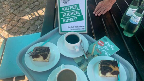 Ein Tablett mit Kaffee und Kuchen und einem Flyer von Kultur für Jeden. auf einem Tisch. Auf dem Tisch stehen zwei Wasserflaschen und ein kleiner Aufsteller mit kleinem Plakat. Text: Keffee, Kuchen,, Kunst! Herzlich willkommen im museums Café.