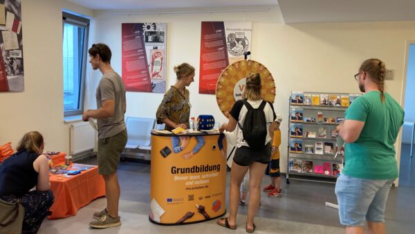 Ein Raum und eine kleiner gelber Stand Text am Stand: "Grundbildung. Besser lesen, schreiben und rechnen."         Ein Kind dreht an einem Glücksrad eine Frau steht am Stand. Wenige weitere Personen sehen sich im Raum um.