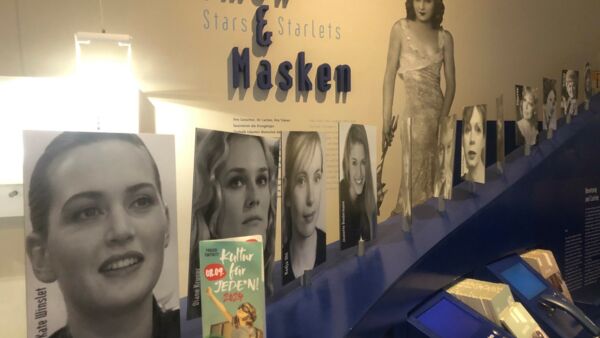 An der Wand steht: "Mimen und Masken - Stars und Starlets" viele schwarz-weiß Fotos von Schauspielerinnen auf einem blauen Bord und ein Flyer von Kultur für Jeden.